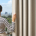 A person parting a beige Sinclair block out curtain to reveal a city skyline view through the window