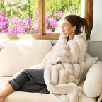 A woman sits on a cream sofa wrapped in a chunky, textured faux fur throw blanket. She is positioned in front of a large window with a view of bright pink flowers outside, wearing dark gray pajama pants.