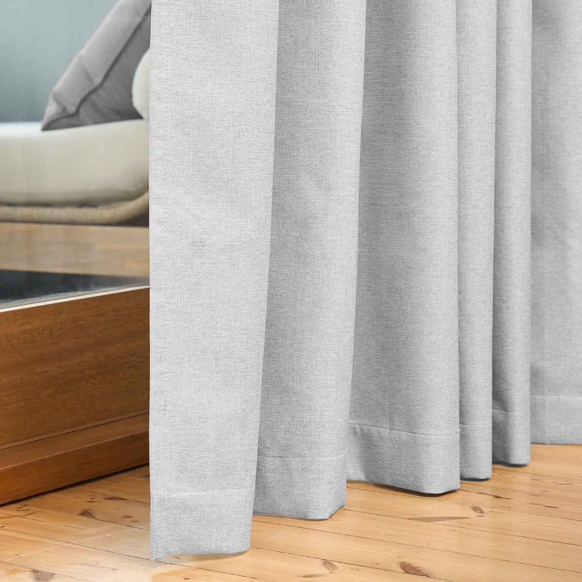 Close-up of the Dakota curtain's hem pooling on a wooden floor in pearl grey, beside a daybed
